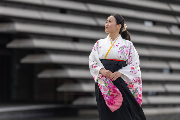 A colour photograph in front of the V&A Dundee you can see the slatted architecture of the museum in the background. A Japanese woman is in the centre of the image looks to the left she wears a kimono which is white on top with pink flowers on the sleeves and has a black skirt.