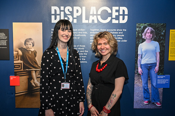 Photograph of two young women standing in front of a display. The woman on the left has dark hard and a black and white spotted top. The woman on the right has a black shirt and red beaded necklace. 