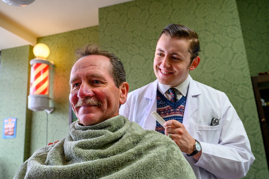 One man sits in a barbers chair with a towel round his neck and shoulders, a hairdresser in a white coat stands behind him. In the back ground there is a red and white barber's shop sign.