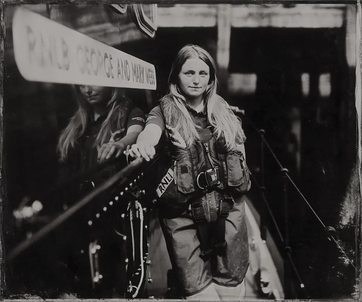 A young woman stands on what could be a RNLI boat, she leans on the boat with her arm outstretched resting on the side of the boat. She looks straight at the camera, she has long blonde hair and wears protective clothing. The image is a black and white photograph. On the side of the boat is a blurred sign which reads 'RNLB George and Mary Webb'