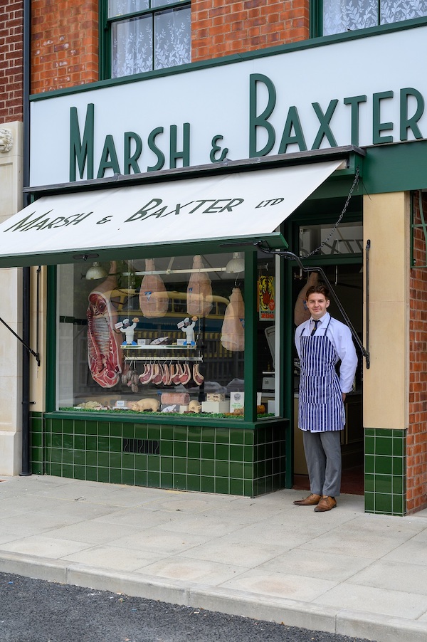 A butchers shop front, with green tiles and a white awning that says Marsh & Baxter, a butcher stands smiling at the entrance in an apron.