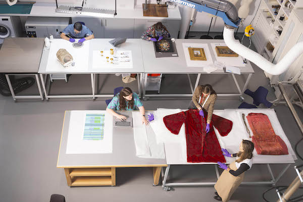 Photograph of a museum conservation studio taken from above. Five people work on museum objects on large desks, four of them wear purple nitrile gloves. 