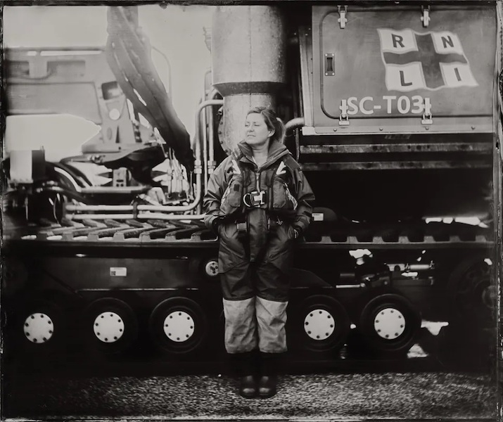 A black and white photograph of a woman standing in front of an RNLI vehicle her body faces forwards but her face looks off to the left of the image. She wears protective clothing and big black boots, her hair is in a pony tail.