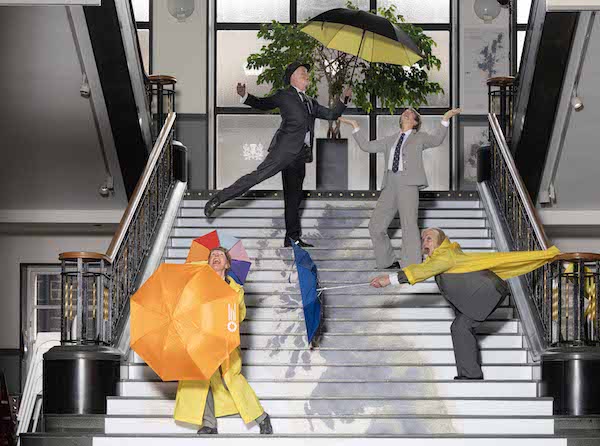 Photograph of a large set of stairs on the stairs are four people posing in raincoats and umbrellas, they have humorous and animated expressions as they look to the sky for rain. 