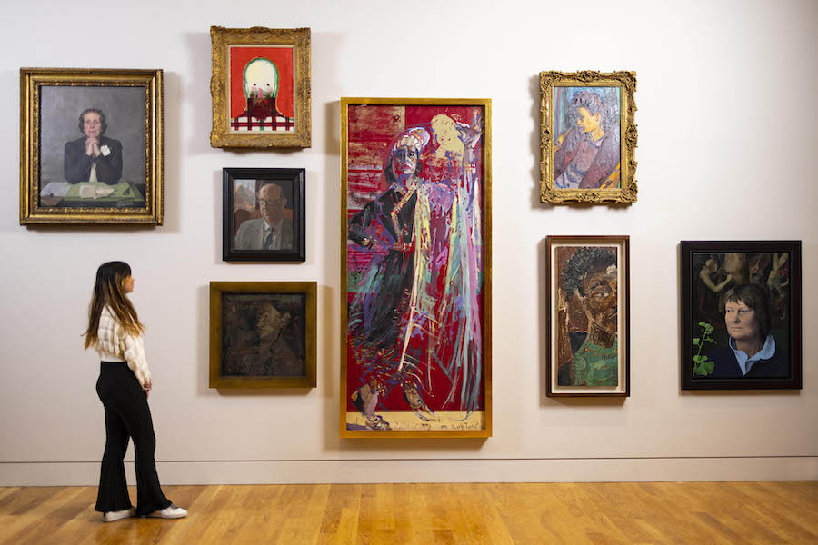 A young woman with long hair, white top and black trousers, stands looking at a gallery wall. There are 8 portraits in different styles on the wall in a cloud hang.