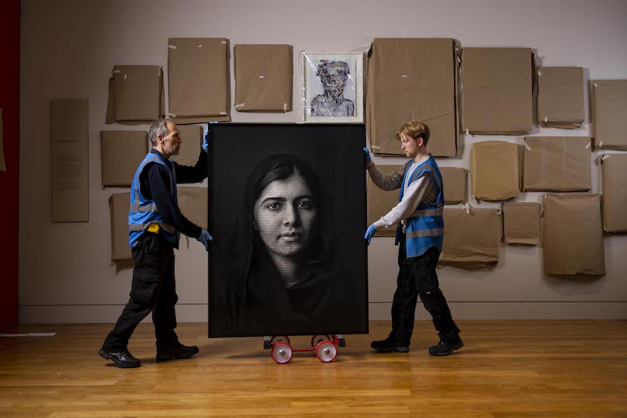 Two workmen carry a portrait of human rights campaigner Malala behind them a gallery wall is covered with paintings each one is covered in brown paper