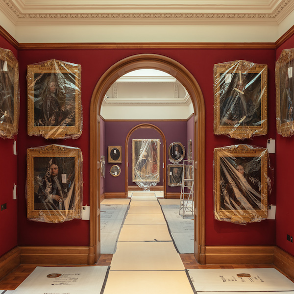 a photograph looking through to the following gallery spaces. The floor is covered in protective material and the paintings on the walls are covered in plastic sheeting. Walls are a deep red and the picture frames are golden 
