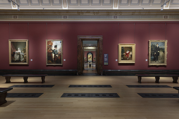 Photograph of the Central Hall at the National Gallery, the walls are a dark red and the floor a light brown, in the centre of the image you can see through to the galleries and painting beyond. On a far wall a painting of a horse. In this gallery either side of the doorway are four large portraits.