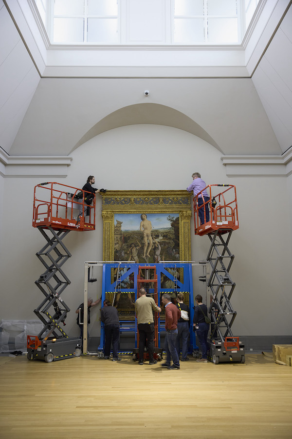 Photograph of the installation of a large painting at the National gallery, two mechanical platforms are extended at the side of the painting with gallery staff in each one. In front of the painting 6 gallery staff use equipment to move the painting into place. 