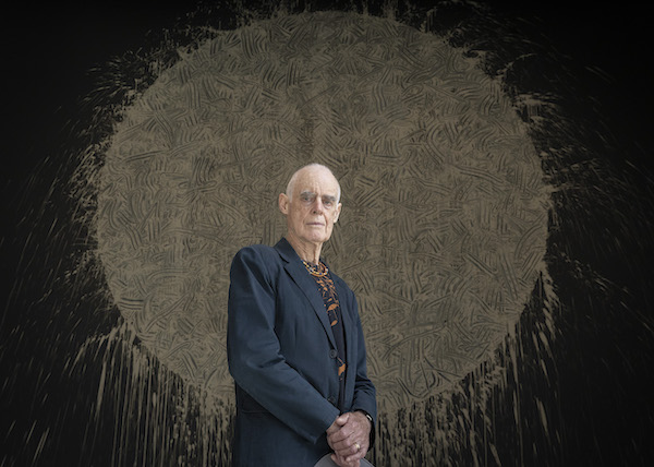 A photograph of the artist Sir Richard Long, a mature white man in a suit with closely cropped white hair. He is standing hands folded in front of him staring at the camera. Behind him is his artwork a large brown circle with markings, the brown is made of mud and the circle splays out at the edges. 