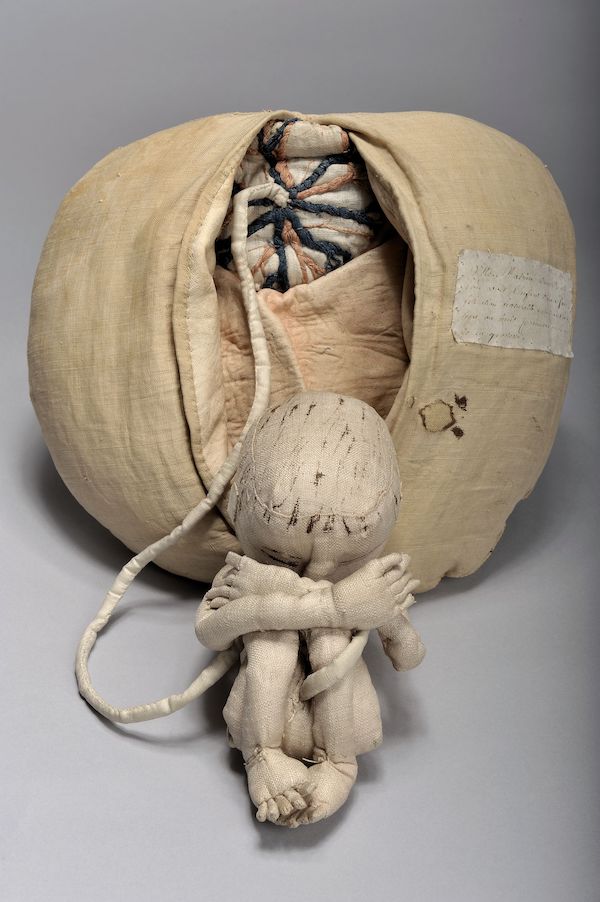 The photograph is of a fabric womb an umbilical chord comes out and is attached to baby curled up in front. The mannequin was used as a teaching tool for midwives. 