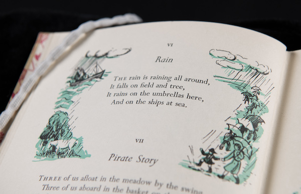 Book of poems open at the page of a poem entitled Rain. The poem is surrounded by an illustration of rain and umbrellas. 