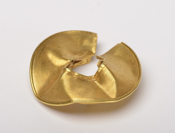 Photograph of a gold lock ring. Circular in shape it has a whole in the middle of the disc. It is damaged and dented.  