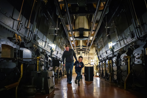 Photograph of the inside of the engine room of the ship HMS Warrior. A man and young child are shown, the child is running towards the camera.