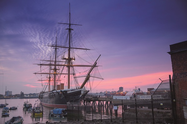 Photograph of a ship with sails in harbour. The sky shows a sunset of purples and pinks. 