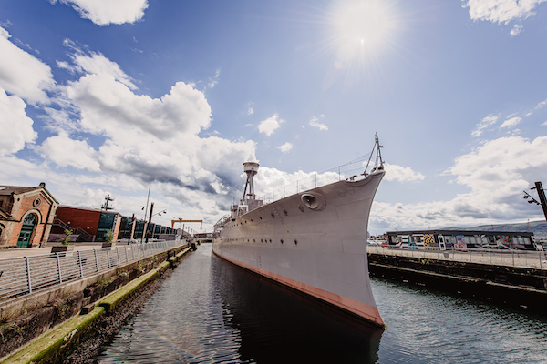Photograph of a ship in a dock. The view shows the bow of the ship and the sky is a light blue with white clouds.
