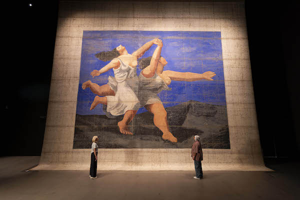 Photograph of a large cloth hanging which was used on a theatre stage. It shows two women running arms extended, hair flying behind them. They are barefoot in white dresses with their left breast bared. It has a blue sky background.