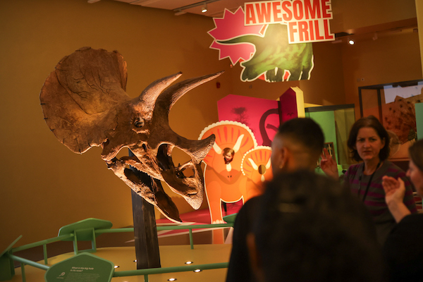 Photograph of visitors at a museum exhibition. In the background is a large Triceratops skull and a bright red sign that says Awesome Frill