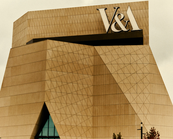 Photography of a building on the top are the initials V&A East the building has geometric shapes scored into the surface. It is has an anguler sloped exterior.