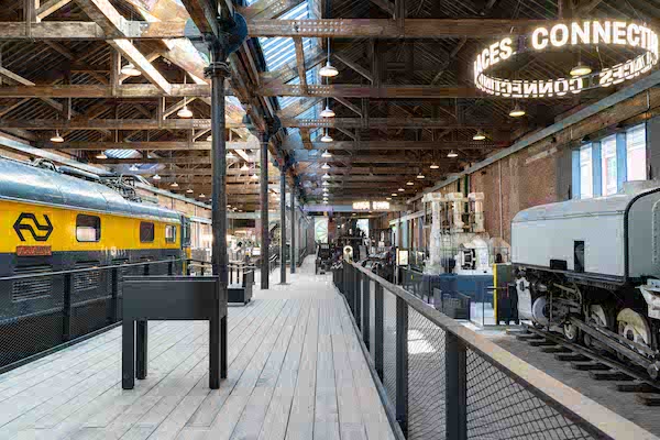 Photograph of a large museum exhibition space, there is a wooden floor stretching into the distance with a train to the left and a small train carriage to the right. 