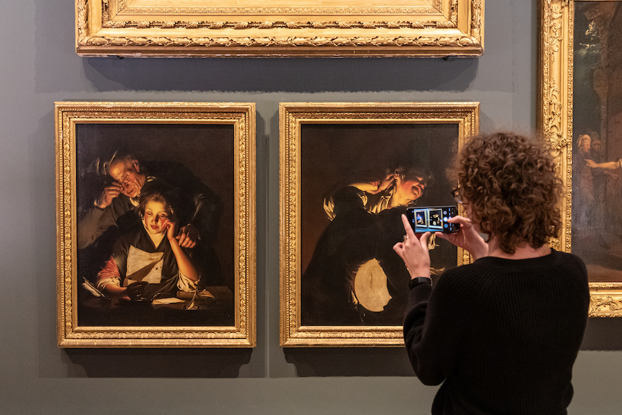 A visitor stands before two Joseph Wright oil paintings she holds up her phone to take a picture