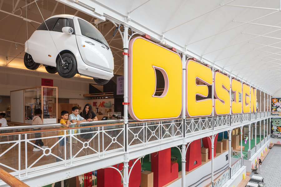 View of the design gallery, the word Design is spelled out in large yellow letters on the side of the gallery there is a white small electric car suspended from the ceiling